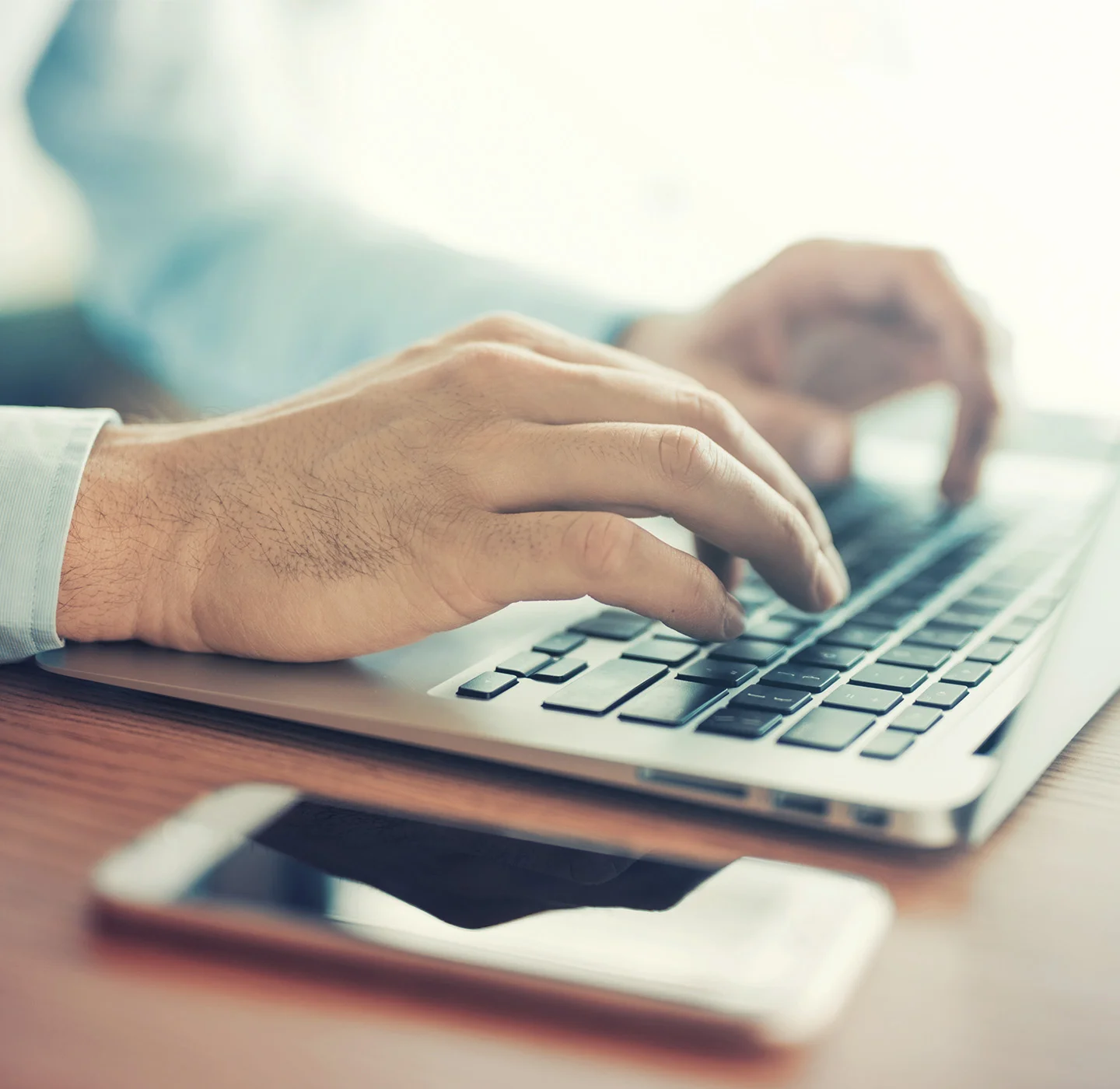 Close-up at hands writing on laptop, with his phone laying beside the computer. Showing different ways to contact our Support